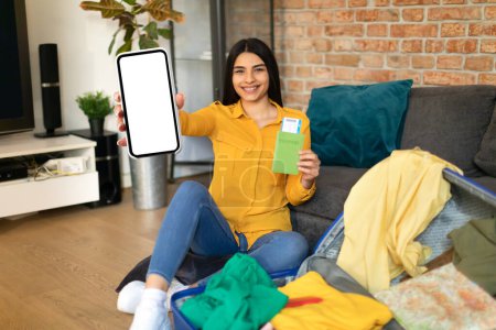 Photo for Booking app concept. Happy spanish tourist lady showing big blank cellphone while packing luggage at home and holding passport with tickets, ready for vacation trip, mockup - Royalty Free Image