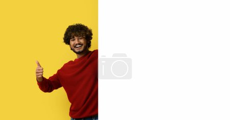 Téléchargez les photos : Joyeux beau jeune homme indien bouclé dans une tenue décontractée élégante posant par une énorme plaque blanche publicitaire avec maquette, montrant pouce vers le haut sur fond de studio jaune, comme une grande offre, espace de copie - en image libre de droit
