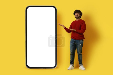 Photo for Nice offer. Smiling handsome young indian guy with curly hair in casual outfit hipster posing over yellow studio background, pointing at big phone with blank screen, full length, mockup - Royalty Free Image