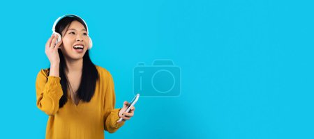 Téléchargez les photos : Joyeuse femme asiatique millénaire heureuse dans l'écoute occasionnelle de la musique, en utilisant des écouteurs stéréo sans fil modernes et smartphone, en regardant l'espace de copie, fond de studio bleu, application musicale divertissante - en image libre de droit