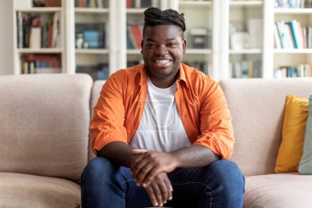 Téléchargez les photos : Portrait d'heureux beau jeune homme noir en tenue décontractée avec des tresses assises sur le canapé à la maison, souriant à la caméra, intérieur du salon, espace de copie. Concept de mode de vie des milléniaux - en image libre de droit