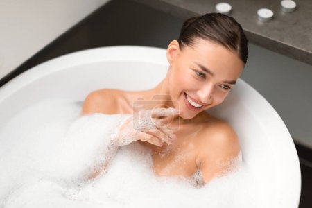 Photo for Young Woman Bathing Lying In Water With Foam Relaxing In White Bathtub In Modern Bathroom Indoors, Above View Shot. Female Taking Bath For Relaxation. Beauty And Wellness - Royalty Free Image