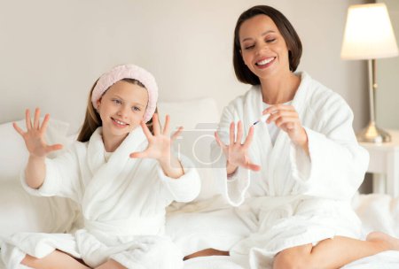 Photo for Glad young caucasian female, small girl in bathrobe sit on white soft bed, do manicure with nail polish, enjoy beauty care in bedroom interior. Weekend mom and daughter, relationship at home - Royalty Free Image