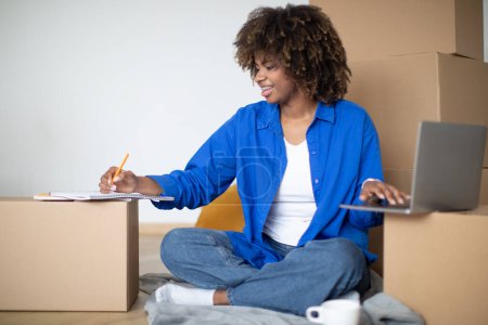 Photo for Happy Black Woman Making Checklist Of Necessities And Using Laptop After Moving Home, Young African American Lady Planning Shopping Or Calculating Budget For House Renovation, Sitting Among Boxes - Royalty Free Image