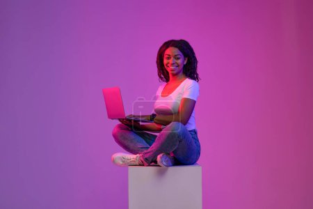 Téléchargez les photos : Heureuse femme noire avec ordinateur portable assis sur le gros cube dans l'éclairage au néon, Souriant jeune femme afro-américaine étude ou de travail en ligne, posant sur dégradé fond violet dans Studio, Copier l'espace - en image libre de droit