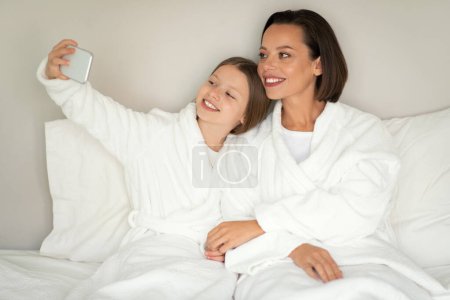 Foto de Sonriente joven mujer, pequeña chica caucásica en albornoz tomar selfie en el teléfono, grabar vídeo para el blog, relajarse en la cama blanda blanca, disfrutar de tiempo libre en el interior del dormitorio. Madre hija relación en casa - Imagen libre de derechos