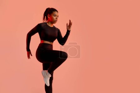 Photo for Athletic african american lady warming up before running, training on peach neon studio background, copy space. Fitness lady standing doing elbow to knee crunch. Sport motivation concept - Royalty Free Image