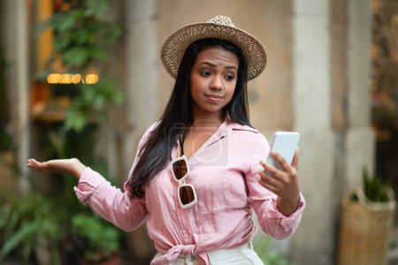 Photo for Confused sad young black female in casual looking at phone has problems in trip, bad internet, mistake in map in city, outside. Device, app for vacation and travel at summer, blog - Royalty Free Image