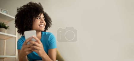 Photo for Happy young curly african american lady typing on smartphone, enjoy social media chat, look at free space in living room interior, panorama. Device, app, ad and offer, surfing in internet at home - Royalty Free Image