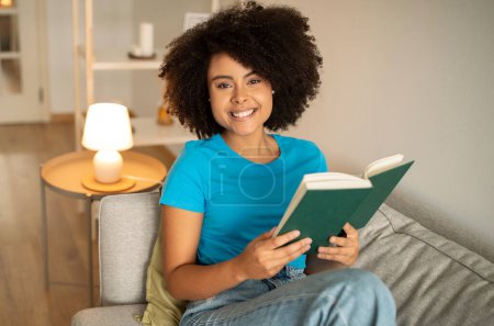 Foto de Feliz joven rizado afroamericano femenino libro de lectura, sentarse en el sofá, disfrutar de relajarse por la noche en el interior de la sala de estar. Hobby, amor a la literatura, estudio, educación y conocimiento en el hogar - Imagen libre de derechos