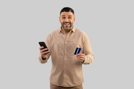 Photo for Excited middle aged man shopping online with mobile phone and credit card, standing on grey studio background, looking at camera with excitement. Businessman making payment via financial application - Royalty Free Image