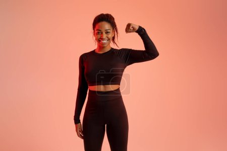 Foto de Tú puedes hacerlo. Retrato de mujer negra emocionada posando en ropa deportiva, mostrando bíceps y sonriendo, de pie sobre fondo de estudio de colores neón. Concepto de deporte y estilo de vida saludable - Imagen libre de derechos