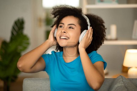 Foto de Sonriente joven rizado mujer afroamericana en auriculares inalámbricos escuchar música en el sofá, disfrutar de vacaciones, se divierte en el interior de la sala de estar. Entretenimiento con canción y aplicación de audio en casa en tiempo libre - Imagen libre de derechos