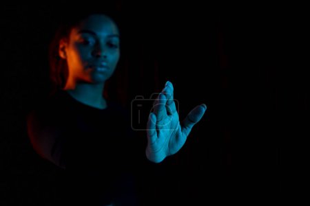 Foto de Concepto de realidad virtual. Joven dama afroamericana estirando la mano y tocando la pantalla táctil invisible, fondo oscuro con luz de neón multicolor, espacio libre - Imagen libre de derechos