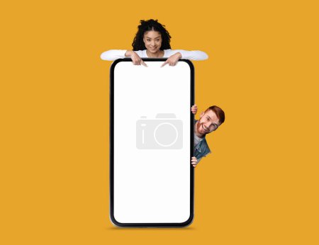 Photo for Online Ad. Cheerful Redhead Man And Black Woman Peeking Out Behind Blank Smartphone With White Screen Isolated Over Yellow Background, Cheerful Couple Recommending New Application Or Website, Mockup - Royalty Free Image