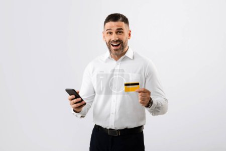 Foto de Emocionado hombre de negocios con camisa usando teléfono inteligente y tarjeta de crédito para ir de compras en línea, pagando por la tan esperada compra en el teléfono móvil, posando sobre un fondo gris - Imagen libre de derechos