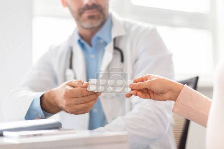 Photo for Middle aged male doctor giving pregnant lady prescribed pills blister pack, sitting at table in clinic office, selective focus on hands holding tablets - Royalty Free Image