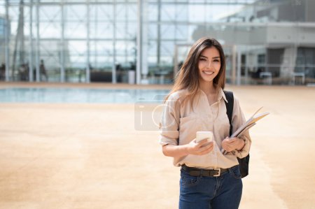 Photo for Smiling young european lady student in casual with backpack, books and smartphone go to lesson at university, outdoor. App for education, study, knowledge in high school, lifestyle with gadget - Royalty Free Image