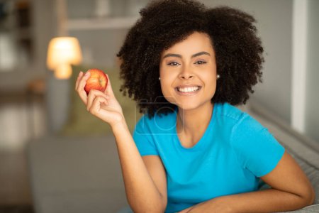 Téléchargez les photos : Souriant jeune femme afro-américaine bouclée avec pomme se trouve sur le canapé, aime se détendre dans l'intérieur du salon. Soins de santé et des dents, nourriture utile, collation et détente à la maison dans le temps libre - en image libre de droit