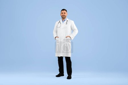 Téléchargez les photos : Médecin professionnel posant portant uniforme blanc tenant les mains dans les poches, regardant vers la caméra, debout sur fond bleu studio, plan complet du médecin généraliste homme - en image libre de droit