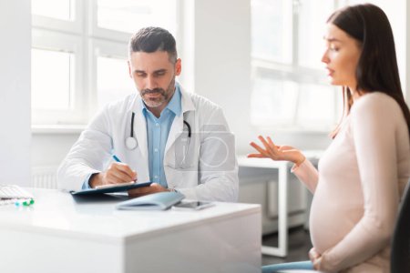 Photo for Young expecting woman visiting doctor middle aged man at private clinic, therapist giving recommendations to pregnant lady. Healthcare during pregnancy concept - Royalty Free Image