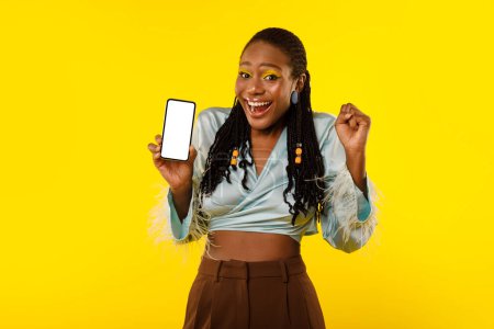 Photo for Great Mobile Offer. Black Female With Bright Makeup Showing Her Smartphone With Empty Screen And Celebrating Luck Shaking Fist Over Yellow Background, Smiling To Camera. Wow App Concept. Mockup - Royalty Free Image