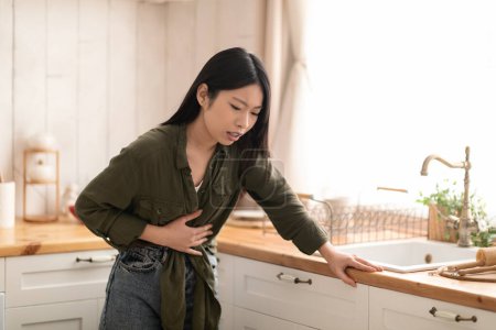 Foto de Mujer asiática joven en casual sintiéndose enferma, tocándose el vientre, de pie junto a la mesa en la cocina, espacio para copiar. Toxicosis durante el embarazo, intoxicación alimentaria, trastornos gastrointestinales concepto - Imagen libre de derechos