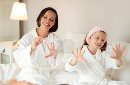 Téléchargez les photos : Joyeux jeune femme caucasienne et petite fille en peignoir s'asseoir sur un lit doux blanc, profiter de manucure avec vernis à ongles et soins de beauté à l'intérieur de la chambre. Mère relation fille à la maison - en image libre de droit