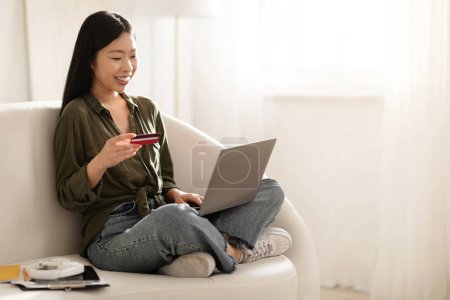 Foto de Sonriente alegre atractiva joven asiática sentada con las piernas en el sofá, el uso de la computadora portátil moderna PC y tarjeta bancaria en casa, el pago de bienes y servicios en Internet, espacio para copiar - Imagen libre de derechos