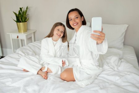 Téléchargez les photos : Heureuse jeune femme caucasienne, adolescente en peignoir prendre selfie au téléphone, tourner la vidéo pour le blog, avoir un appel vidéo, profiter de se détendre sur un lit blanc dans l'intérieur de la chambre. Mère relation fille à la maison - en image libre de droit