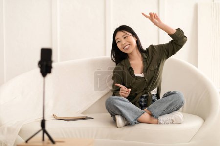 Foto de Lindo bastante elegante millennial asiático lady vlogger grabación de contenido para los seguidores en las redes sociales, sentado en el sofá y el baile, mirando el teléfono celular en el trípode y sonriendo, espacio de copia - Imagen libre de derechos
