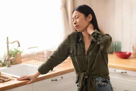 Foto de Mujer asiática joven infeliz en dolor de pie junto al escritorio de la cocina, frotando su cuello, mujer que sufre de tensión muscular del cuello después de incómodo dormir en la mañana, espacio para copiar - Imagen libre de derechos