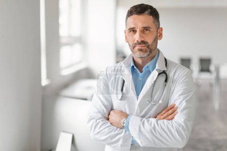 Photo for Healthcare and medicine concept. Portrait of confident middle aged male doctor with folded arms posing and looking at camera, standing in clinic office interior, copy space - Royalty Free Image