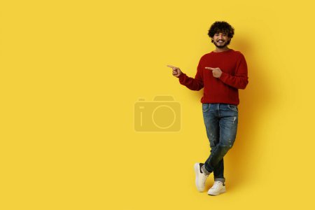 Photo for Nice offer. Cheerful handsome young eastern guy in casual hipster posing alone over yellow studio background, pointing at copy space for advertisement and smiling, full length - Royalty Free Image