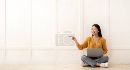 Photo for Cute cheerful pretty young chinese woman in casual sitting on floor with laptop over white blank wall, pointing at copy space and smiling, showing nice online offer, panorama - Royalty Free Image