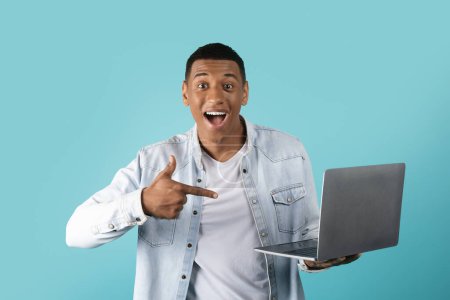 Photo for Glad millennial black male in casual showing finger at laptop, enjoy win isolated on blue studio background. Device presentation, great offer, ad, good news and human emotions - Royalty Free Image