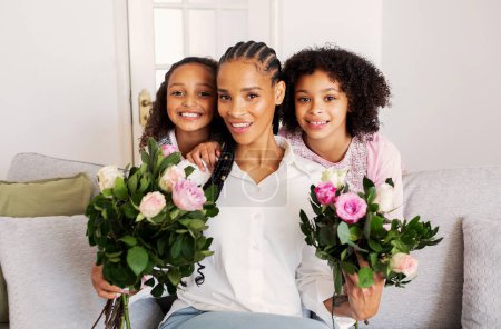 Photo for Cheerful Black Mother And Daughters Posing With Flowers Bouquets Smiling Looking At Camera, Kids Congratulating Mommy On Birthday Celebrating Holiday Sitting On Sofa At Home - Royalty Free Image
