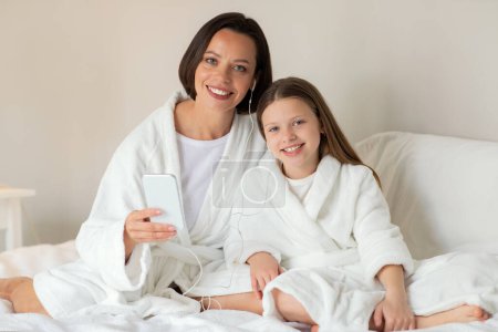 Téléchargez les photos : Petite fille caucasienne souriante, jeune femme en peignoir assis sur un lit blanc avec écouteurs et téléphone, écouter de la musique profiter de temps libre et de soins de beauté dans l'intérieur de la chambre. Maman et fille à la maison - en image libre de droit