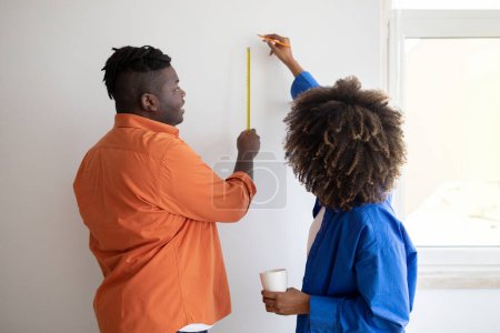 Photo for Black young couple with tape ruler taking measurements on white wall indoors, happy african american spouses doing improvement and renovation at home, planning design in new apartment, rear view - Royalty Free Image