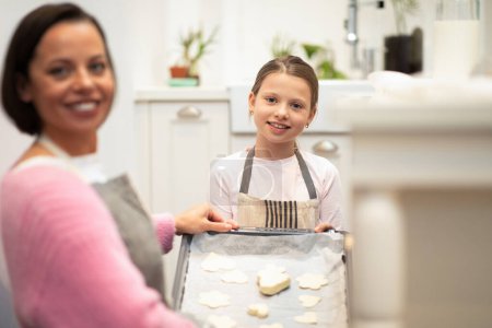 Téléchargez les photos : Joyeux caucasien jeune maman et petite fille dans des tabliers font des biscuits à partir de pâte sur plaque de cuisson à l'intérieur de la cuisine, floue. Boulangerie, cuisine maison, confiserie à la maison - en image libre de droit
