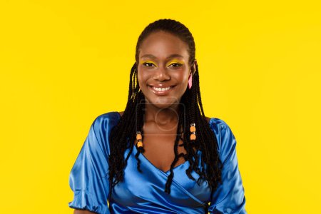 Téléchargez les photos : Portrait de jolie dame afro-américaine tressée avec un maquillage lumineux posant souriant à la caméra sur fond de studio jaune, portant une blouse bleue. Beauté féminine et concept de style - en image libre de droit