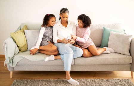 Photo for Happy Black Mother And Two Daughters Using Cellphone And New Mobile Application Sitting On Sofa At Home. Family Of Three Browsing Internet Via Phone. Gadgets And Technology Concept - Royalty Free Image