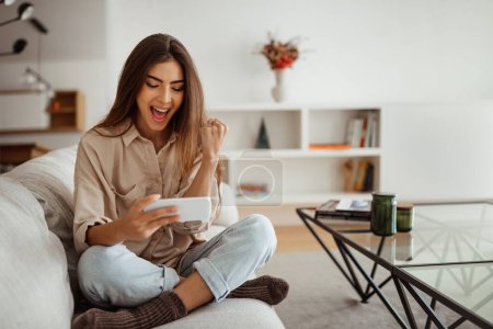 Photo for Glad shocked millennial mixed race woman with open mouth looking at phone, reading message, chatting, enjoy free time in room interior. App for communication remotely, good news at home - Royalty Free Image
