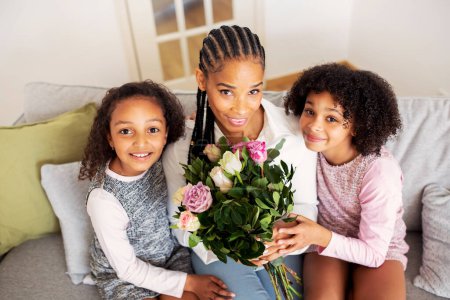 Foto de Feliz madre negra sosteniendo flores recibiendo saludos de hijas preadolescentes, posando sentadas en el sofá y sonriendo a la cámara en casa. Vacaciones en familia, Concepto de Celebración del Día de las Madres - Imagen libre de derechos