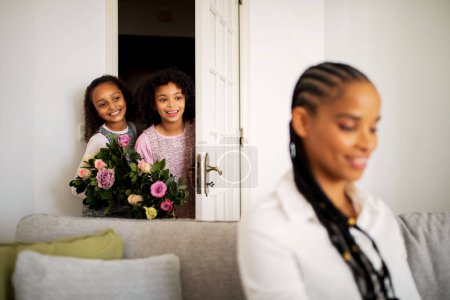 Foto de Día de las Madres. Feliz afroamericano niñas sosteniendo flores de pie detrás de las mamás espalda sorprendente y felicitándola en vacaciones en casa. Enfoque selectivo en los niños - Imagen libre de derechos