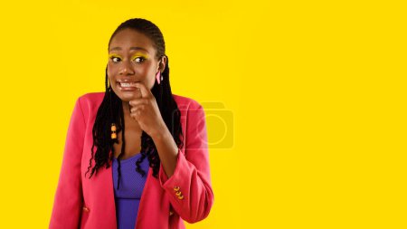 Téléchargez les photos : Dame afro-américaine incertaine pensant tenant doigt près de la bouche exprimant inquiétude et anxiété posant regardant à côté debout sur fond jaune studio. Panorama avec espace vide - en image libre de droit