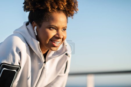 Photo for Smiling Young Black Woman Wearing Earphones And Armband Taking Break While Jogging Outdoors, Closeup Portrait Of Motivated African American Lady Resting After Outside Workout, Copy Space - Royalty Free Image