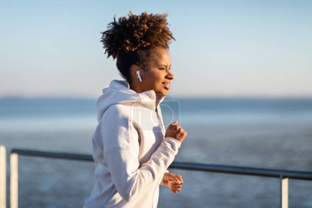 Téléchargez les photos : Bonjour aux sportifs. Souriant noir femme jogging extérieur près de la mer, heureux jeune afro-américaine femme portant des écouteurs sans fil courir dehors et profiter de l'écoute de musique préférée, Copier l'espace - en image libre de droit