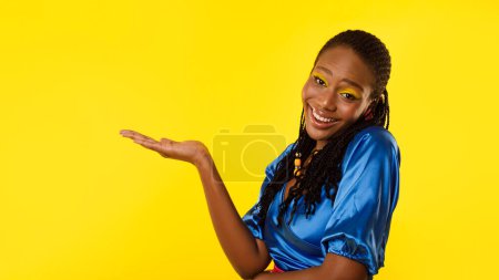 Téléchargez les photos : Joyeuse dame noire tenant la main produit de beauté invisible ou montrant l'espace de copie souriant à la caméra posant sur fond jaune studio. Stylistes de mode Offre Concept. Panorama - en image libre de droit
