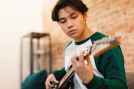 Foto de Japonés adolescente chico jugando guitarra eléctrica tocando cuerdas tomando acordes sentado en sofá en casa. Boy Learning To Play Music Writing New Song On Weekend (en inglés). Hobby y talento. Enfoque selectivo - Imagen libre de derechos